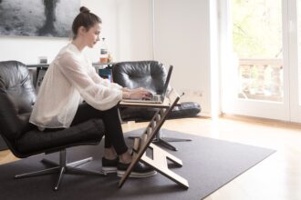Person sitzt ergonomisch an einem höhenverstellbaren Schreibtisch mit Bürostuhl, Monitor auf Augenhöhe und geordnetem Arbeitsplatz.