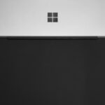 Ein schwarzes Microsoft Surface Dock 2 mit angeschlossenen Kabeln für Monitore und Peripherie auf einem aufgeräumten Schreibtisch.